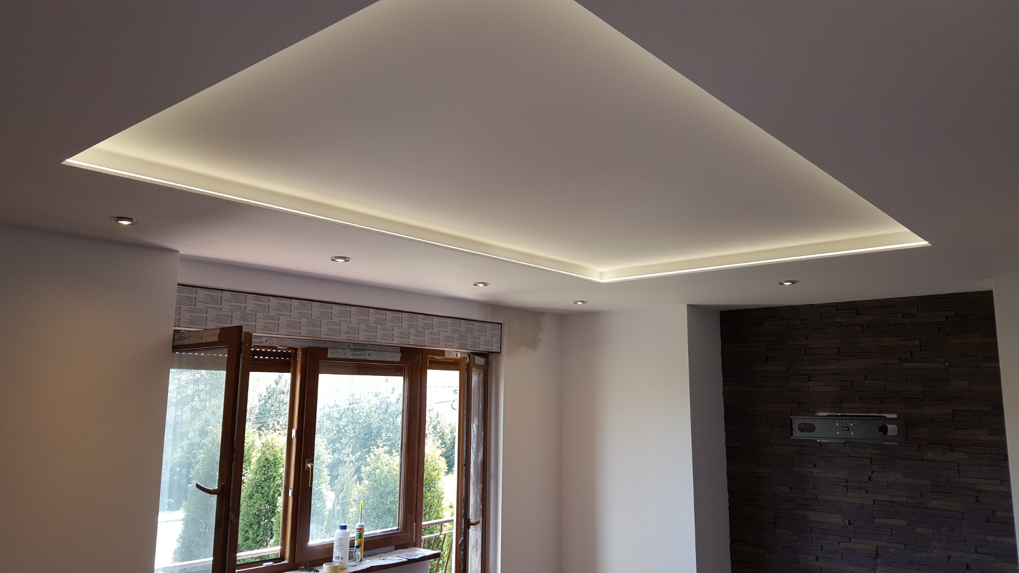 Nowoczesny salon z podwieszanym sufitem z oświetleniem LED, widok na okno z roletami i dekoracyjną ścianę z kamienia.