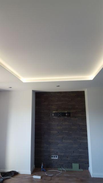 Nowoczesne wnętrze z podświetlanym sufitem napinanym, ściana z dekoracyjnej, ciemnej cegły, widoczne gniazdka elektryczne i przewody na podłodze.