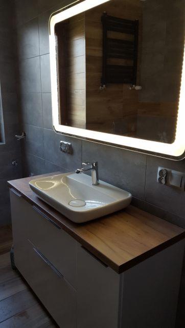 Nowoczesna łazienka z umywalką nablatową na szafce z drewnianym blatem, podświetlane lustro i szare płytki na ścianach.