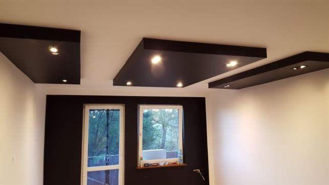 Nowoczesny salon z czarnymi podwieszanymi panelami sufitowymi z punktowym oświetleniem LED, kontrastującymi z białymi ścianami i czarną ścianą z oknem.