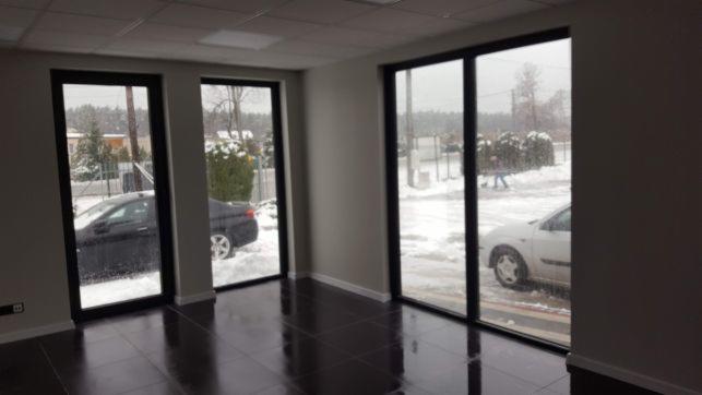 Wnętrze biura z ciemną, błyszczącą podłogą z płytek, trzema dużymi oknami w czarnych ramach, za którymi widać ośnieżony krajobraz i zaparkowane samochody.