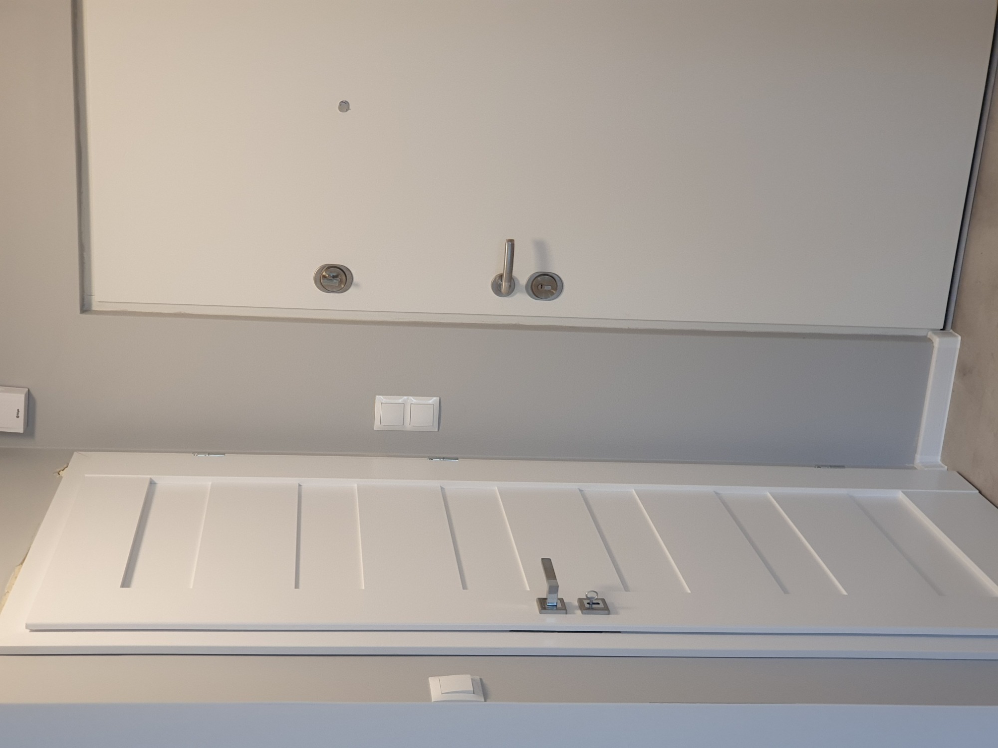 Dwa białe, nowoczesne drzwi wewnętrzne z srebrnymi klamkami i zamkami, umieszczone w szarym pomieszczeniu. Górne drzwi gładkie, dolne z frezowanymi pionowymi liniami.