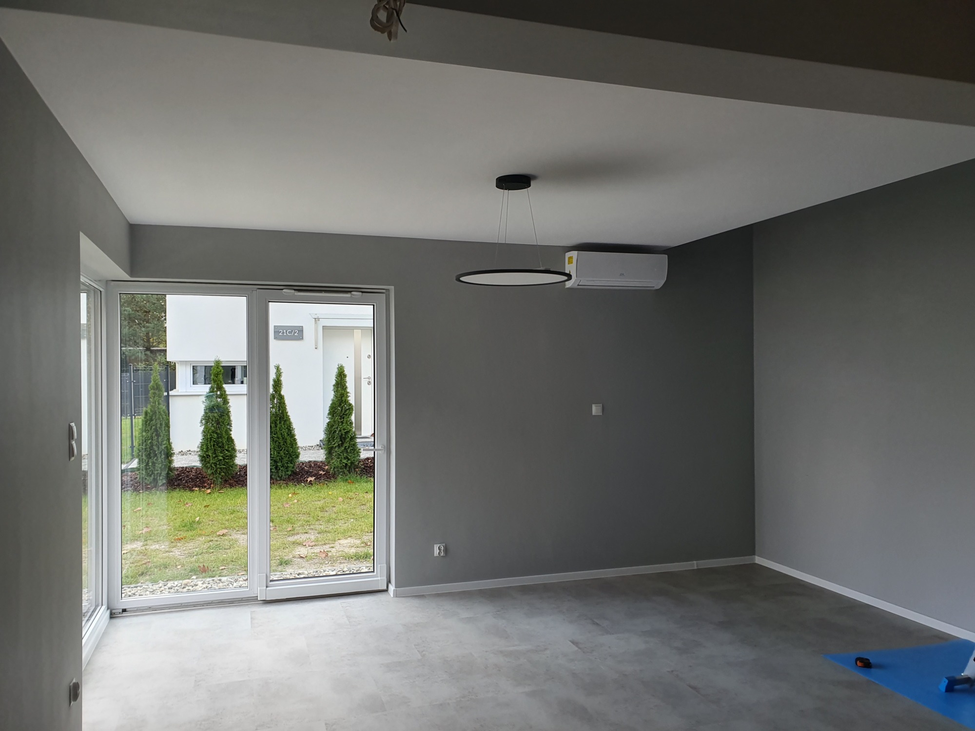 Wnętrze pokoju po malowaniu ścian na szaro, z widokiem na ogród przez przeszklone drzwi przesuwne i nowoczesną lampą sufitową.