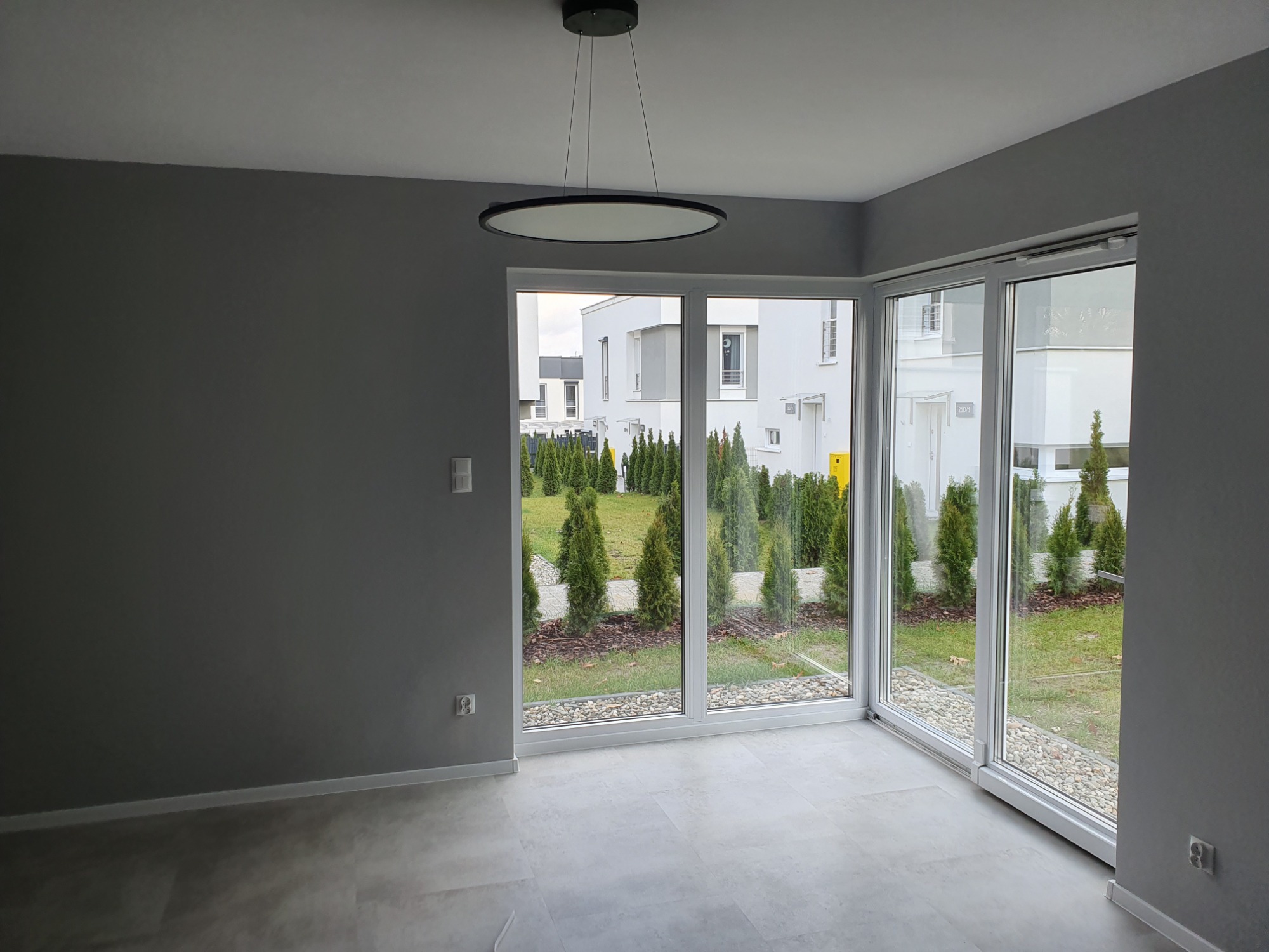Pusty pokój z szarymi ścianami i panelami podłogowymi w odcieniach szarości, duże okna z widokiem na ogród z tujami i nowoczesne budynki mieszkalne, okrągła lampa sufitowa.