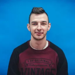 Portret młodego mężczyzny na tle jednolitego, jasnoniebieskiego tła. Ma ciemne włosy zaczesane do góry i ubrany jest w bordowo-czarną bluzę z napisem 'Vintage'.