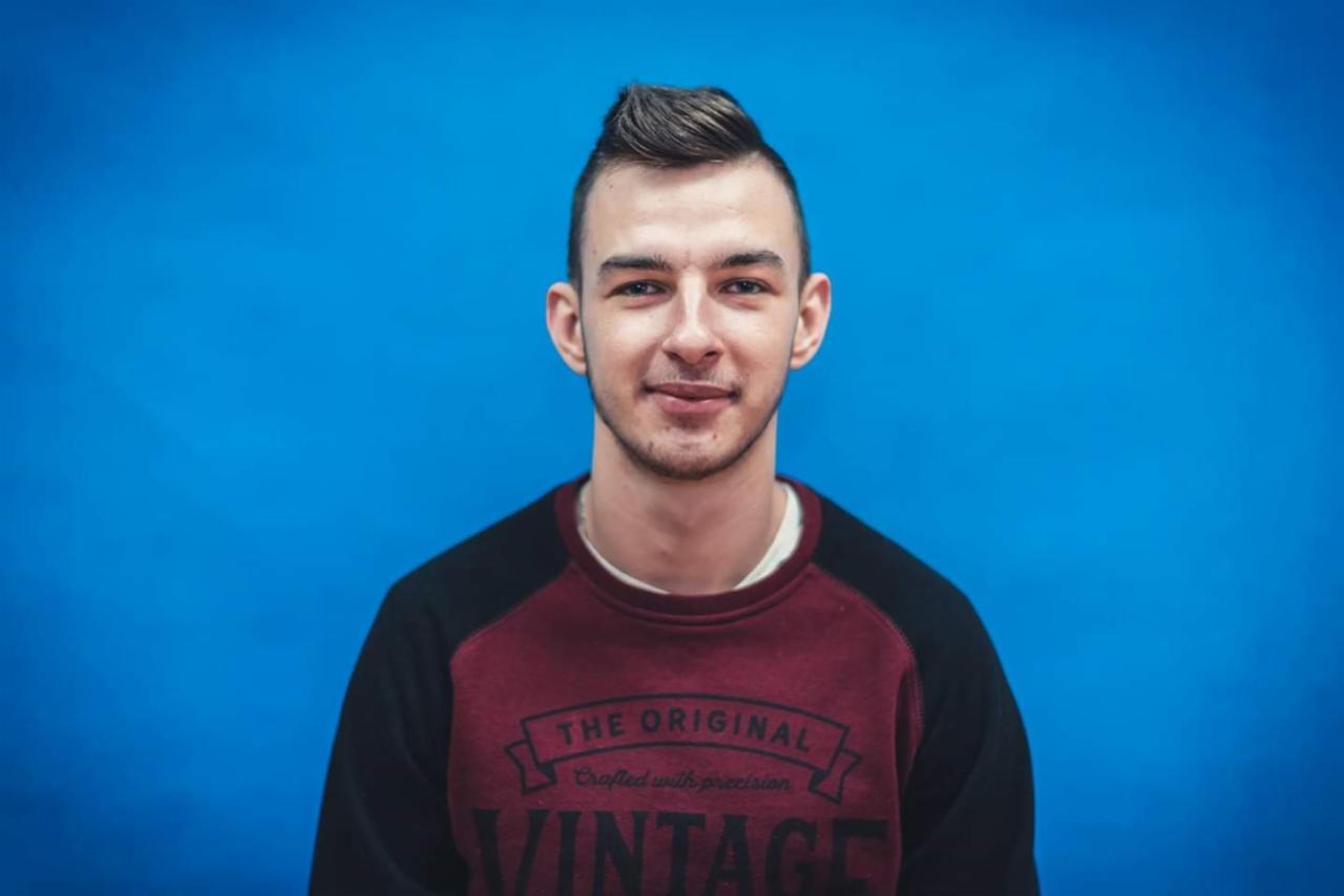 Portret młodego mężczyzny na tle jednolitego, jasnoniebieskiego tła. Ma ciemne włosy zaczesane do góry i ubrany jest w bordowo-czarną bluzę z napisem 'Vintage'.