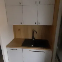 Nowoczesna, mała kuchnia z białymi szafkami górnymi i dolnymi, drewnianym blatem i czarnym zlewozmywakiem z baterią. Widoczna część podłogi z jasnymi panelami. Oświetlenie górne jest słabe.