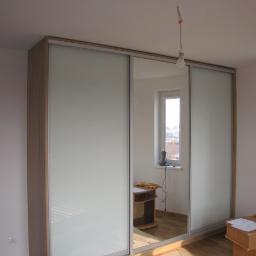 Piofen Piotr Bryll - Nowoczesna szafa z przesuwnymi, mlecznymi drzwiami, częściowo zamontowana, z widoczną instalacją elektryczną i narzędziami na podłodze, odbicie pokoju w lustrzanej części drzwi.