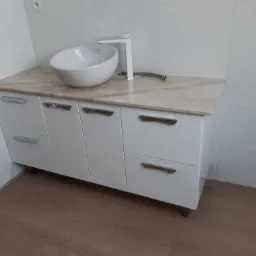 Biała szafka łazienkowa z umywalką nablatową i marmurowym blatem, nowoczesna armatura, widoczny fragment wężyka doprowadzającego wodę.