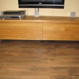 Piofen Piotr Bryll - Drewniana szafka RTV z dwoma drzwiczkami, na której stoi telewizor, odtwarzacz DVD i głośniki, na brązowej podłodze.