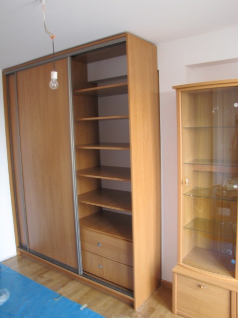 Jasna, drewniana szafa z przesuwnymi drzwiami, z otwartą częścią regałową i szufladami, obok witryna, na podłodze folia zabezpieczająca, w tle surowa żarówka na kablu.