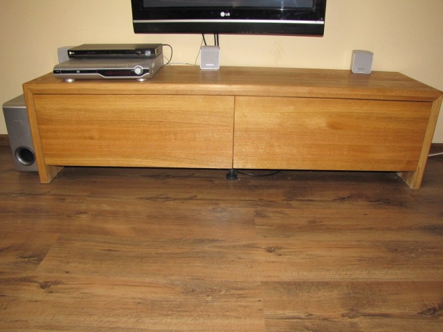 Drewniana szafka RTV z dwoma drzwiczkami, na której stoi telewizor, odtwarzacz DVD i głośniki, na brązowej podłodze.