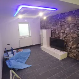 Pokój po remoncie z szarymi płytkami podłogowymi, ścianą z imitacją kamienia, podwieszanym sufitem z niebieskim oświetleniem LED, zabezpieczonymi meblami i widocznymi śladami prac wykończeniowych.