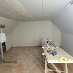Poddasze w trakcie zabudowy płytami kartonowo-gipsowymi, widoczne narzędzia i laser krzyżowy na prowizorycznym stole roboczym, surowe wykończenie z widocznymi łączeniami płyt.