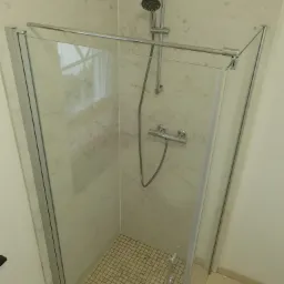 Kabina prysznicowa z transparentną szybą i chromowanymi profilami, wykończona marmurowymi płytami, z drobną mozaiką na podłodze brodzika i odpływem liniowym.