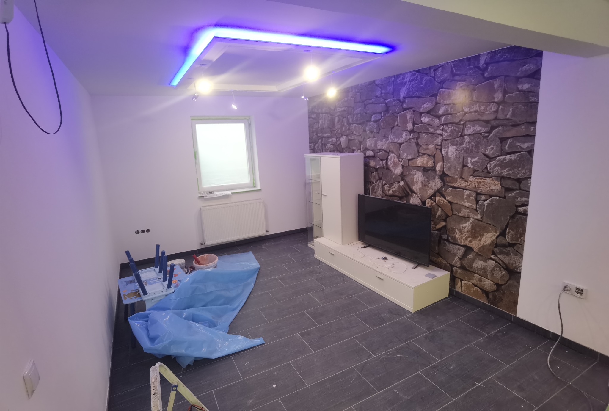 Pokój po remoncie z szarymi płytkami podłogowymi, ścianą z imitacją kamienia, podwieszanym sufitem z niebieskim oświetleniem LED, zabezpieczonymi meblami i widocznymi śladami prac wykończeniowych.