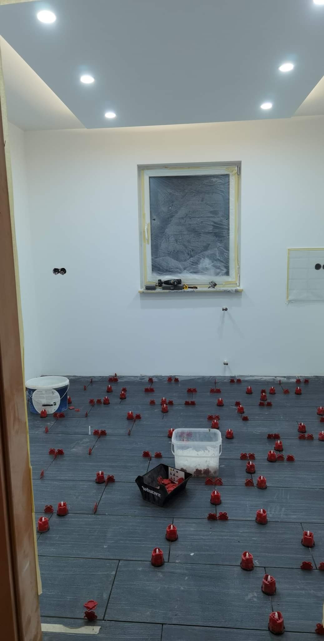 Układanie płytek podłogowych w szarym odcieniu z użyciem czerwonych klipsów poziomujących; widoczne narzędzia i wiadro z zaprawą na zabezpieczonym oknie w tle.