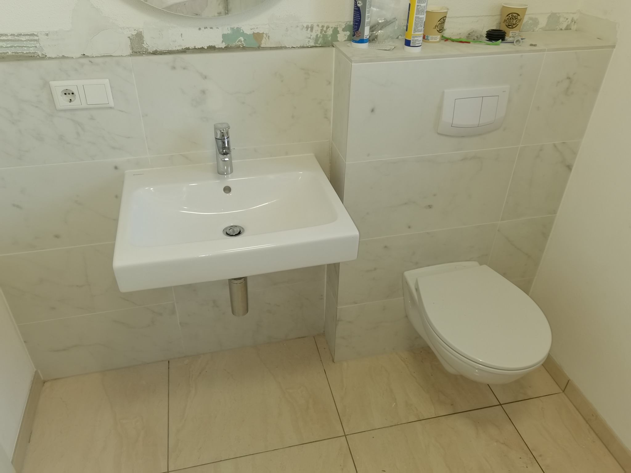 Biała umywalka z baterią i podwieszana toaleta w wykończonej łazience z marmurowymi płytkami, widoczne gniazdko elektryczne i przycisk spłuczki