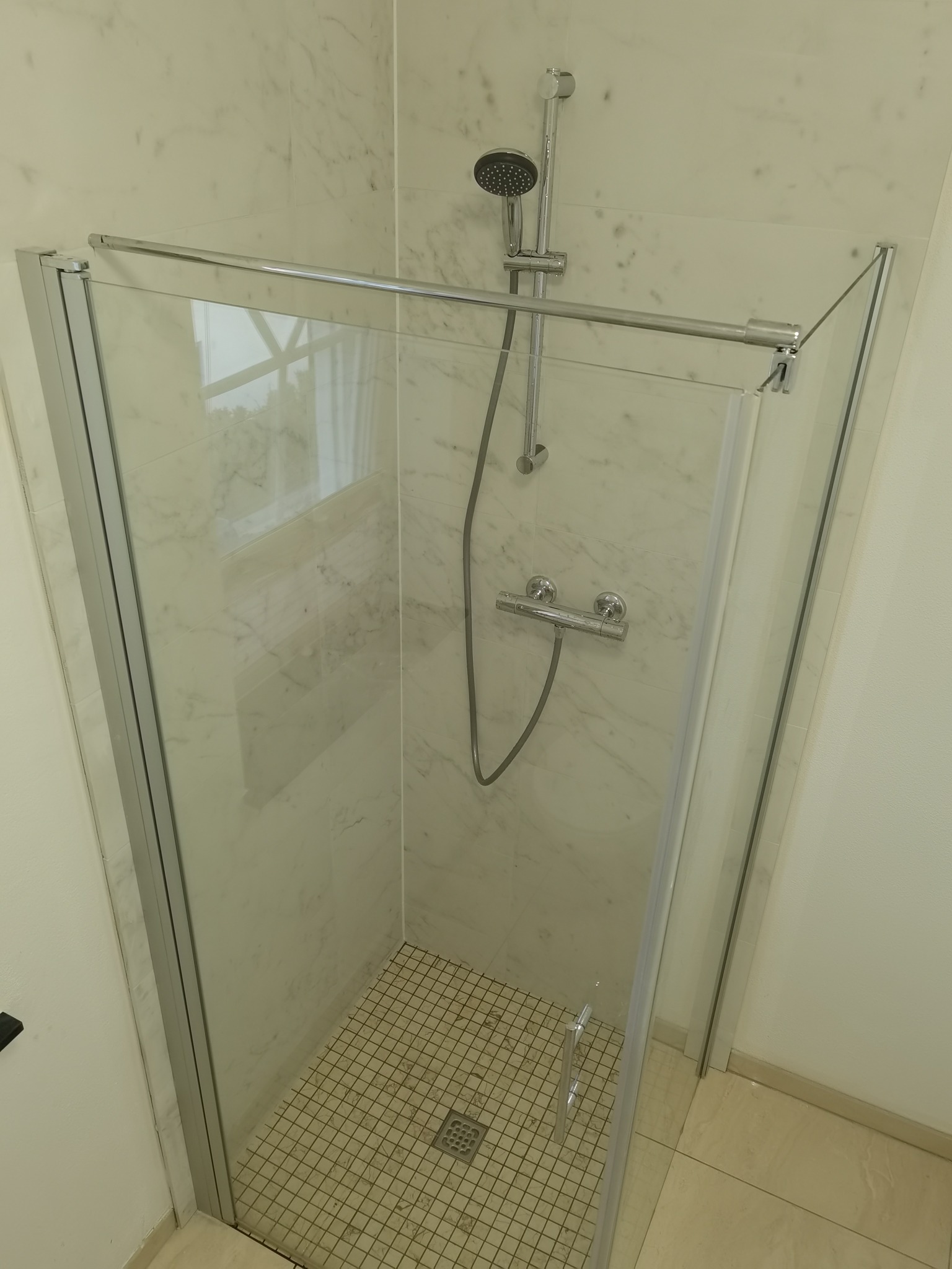 Kabina prysznicowa z transparentną szybą i chromowanymi profilami, wykończona marmurowymi płytami, z drobną mozaiką na podłodze brodzika i odpływem liniowym.