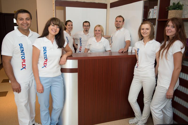 Zespół kliniki dentystycznej DentoPlex w białych uniformach z logo, pozujący za recepcją z drewnianym blatem.
