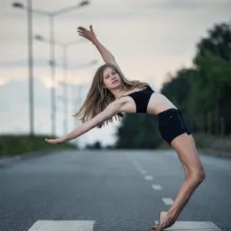 Młoda kobieta w czarnym stroju wykonuje łuk na jezdni z białymi pasami, na tle słupy oświetleniowe i drzewa.