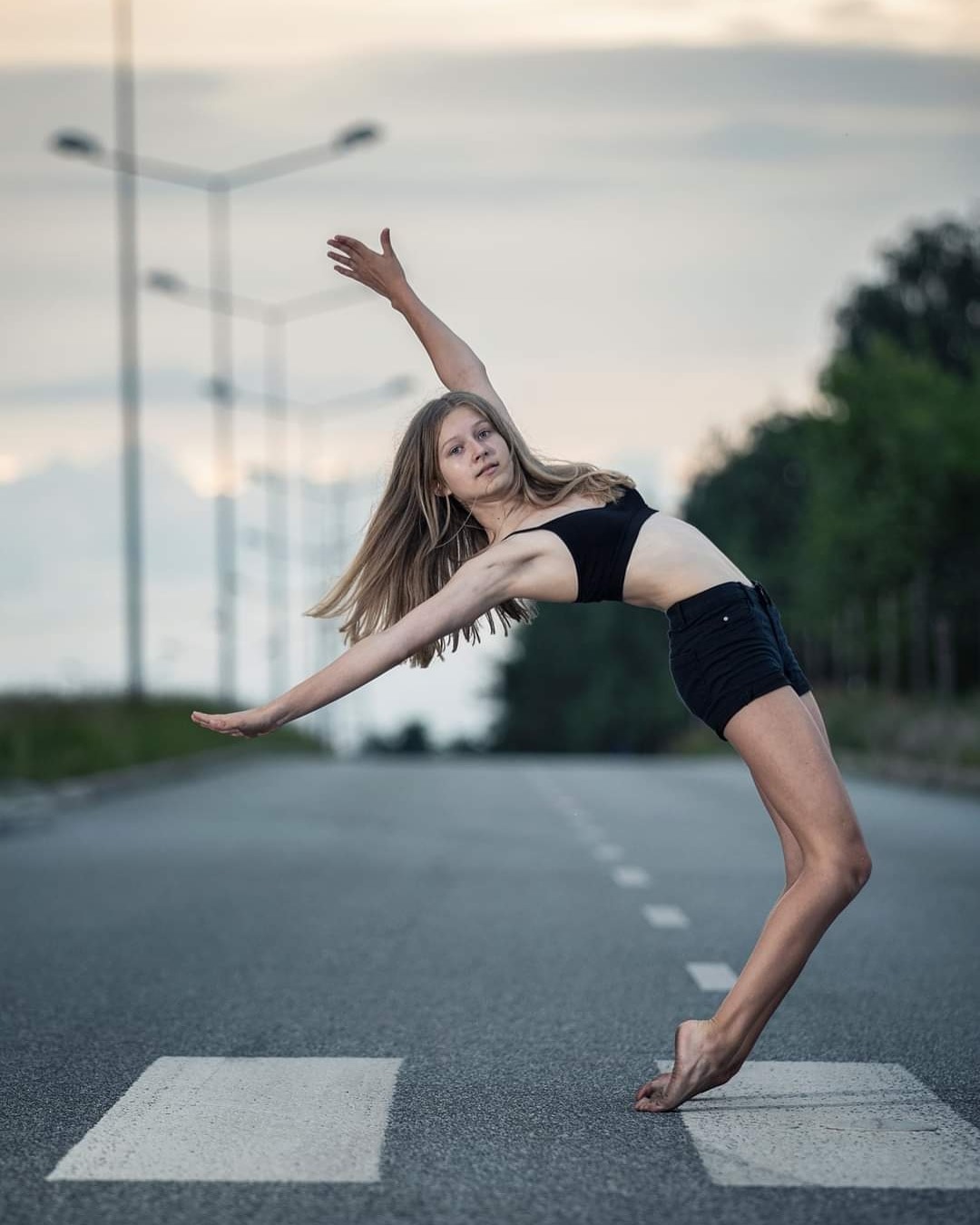 Młoda kobieta w czarnym stroju wykonuje łuk na jezdni z białymi pasami, na tle słupy oświetleniowe i drzewa.