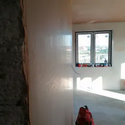 Świeżo wyszpachlowana ściana w trakcie schnięcia, widoczne niedoskonałości i struktura gładzi, okno z widokiem na ogród, czerwona torba narzędziowa na podłodze.