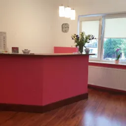 Recepcja w salonie: różowy kontuar z drewnianym blatem, w tle okno z kwiatami, zegar na ścianie i ulotka reklamowa Dr. Grandel.