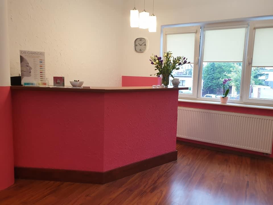 Recepcja w salonie: różowy kontuar z drewnianym blatem, w tle okno z kwiatami, zegar na ścianie i ulotka reklamowa Dr. Grandel.