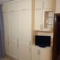 Kremowe szafy z metalowymi uchwytami, jedna z przeszkloną witryną, z telewizorem na blacie, obok zasłona i widoczny fragment zegara na ścianie.