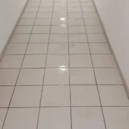 Długi, wąski korytarz wyłożony jasnymi, kwadratowymi płytkami z ciemnymi fugami, świeżo umyty, z widocznymi refleksami światła na powierzchni.