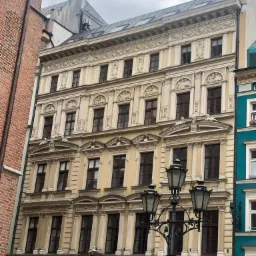 Fasada odnowionej, secesyjnej kamienicy z beżową elewacją, ciemnymi oknami i ozdobnymi detalami architektonicznymi, widoczna z poziomu ulicy, z charakterystycznymi latarniami w pierwszym planie.