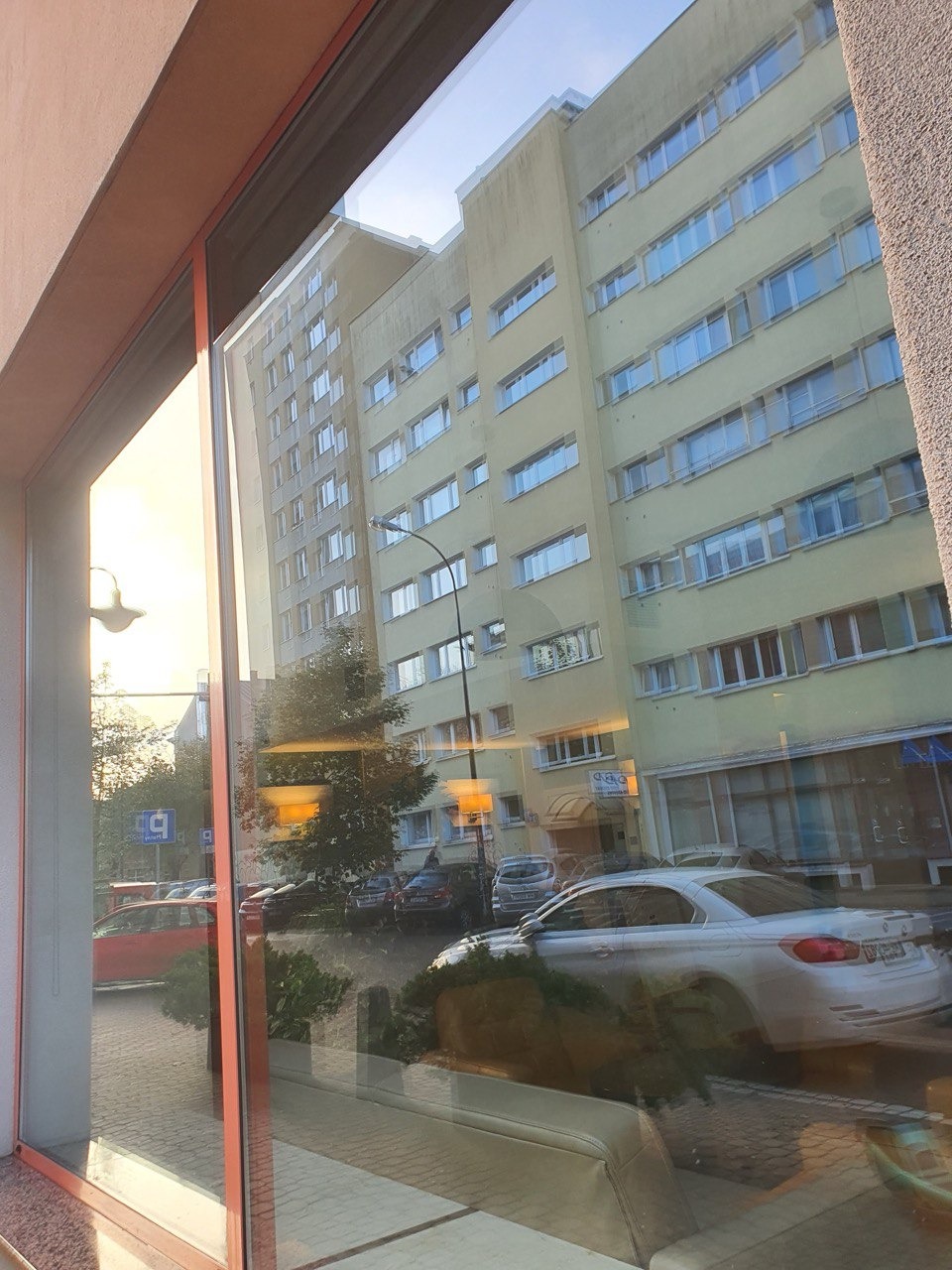 Odbicie miejskiego krajobrazu w czystej szybie okiennej z pomarańczową ramą. Widoczny budynek, drzewa i zaparkowane samochody.