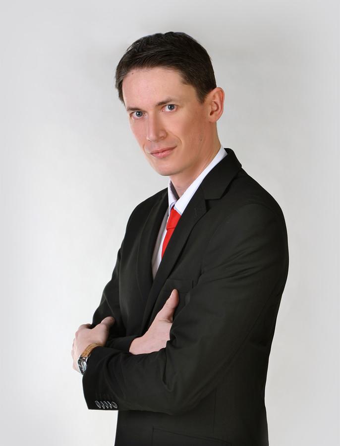 Portret biznesowy mężczyzny w czarnym garniturze i czerwonym krawacie, z założonymi rękami, delikatny uśmiech, jasne tło. Widoczny zegarek na nadgarstku.