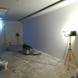 Wnętrze pokoju w trakcie remontu, ściany przygotowane pod malowanie, widoczne narzędzia, wiadro z farbą, szpachla, lampa halogenowa na statywie, odkurzacz przemysłowy.