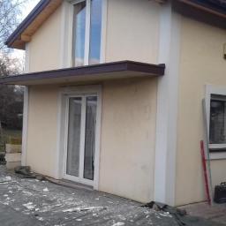 Usługi Remontowo-budowlane Damian Krasnodębski - Dom z oknami po remoncie elewacji, widoczne ślady prac, drabina oparta o ścianę, tło z drzewami bez liści i przyczepą kempingową.