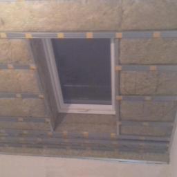 Usługi Remontowo-budowlane Damian Krasnodębski - Montaż izolacji z wełny mineralnej wokół okna dachowego na poddaszu, widoczne metalowe profile i taśmy mocujące.