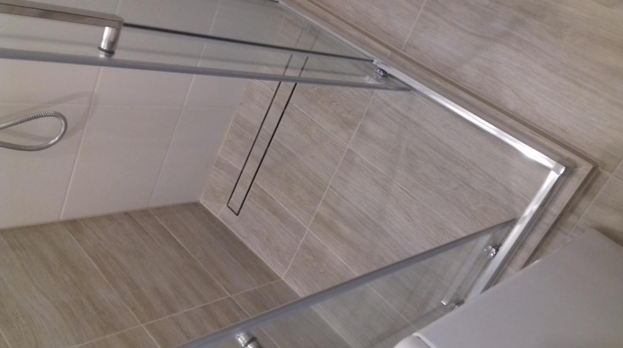 Szklana kabina prysznicowa z odpływem liniowym w podłodze wyłożonej płytkami imitującymi drewno.