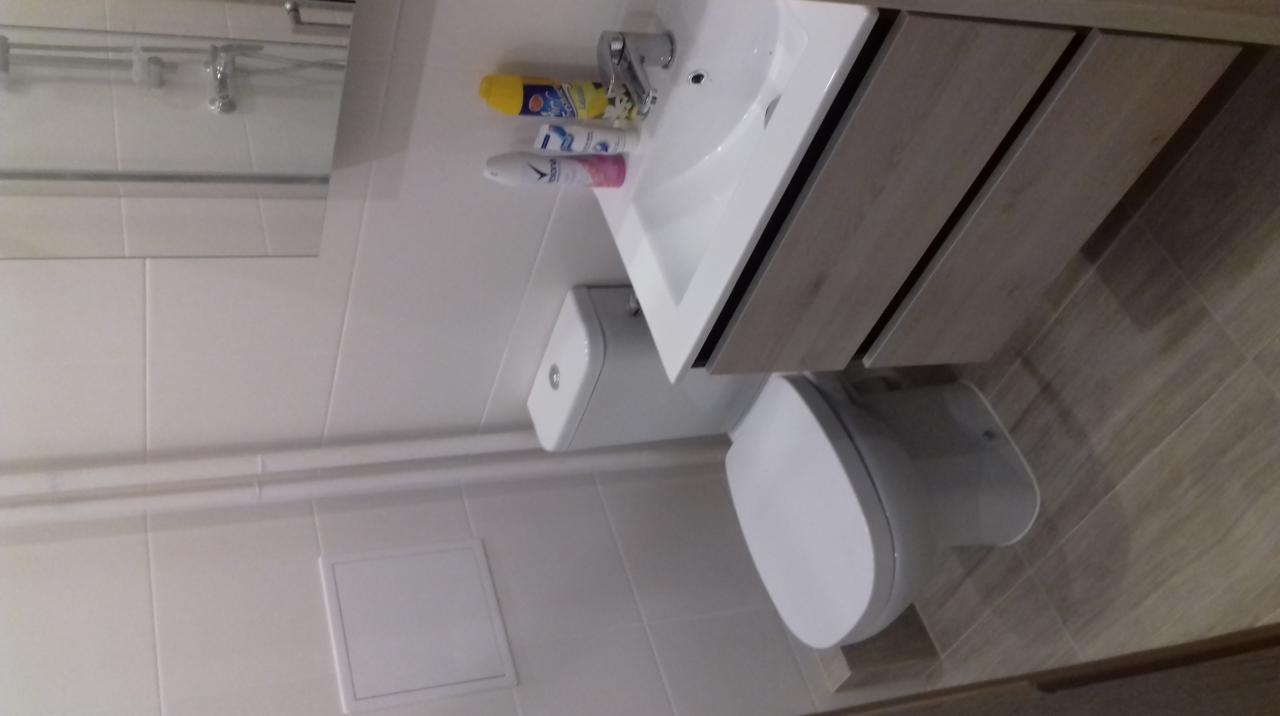 Widok na białą toaletę z deską wolnoopadającą, umywalkę z szafką o drewnianym wykończeniu, baterią i kosmetykami, oraz białe płytki na ścianach w łazience.