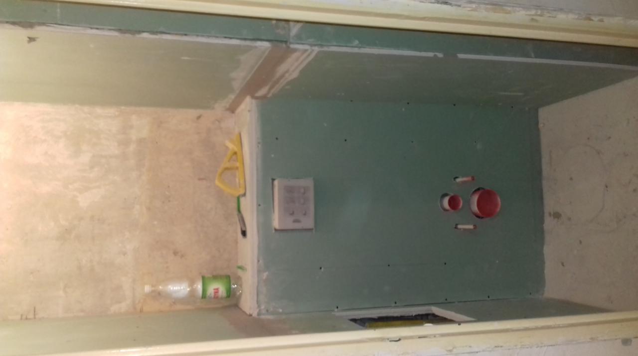 Fragment ściany z zabudową z płyt gipsowo-kartonowych w trakcie prac wykończeniowych, widoczne otwory instalacyjne, żółta szpachelka i butelka wody.