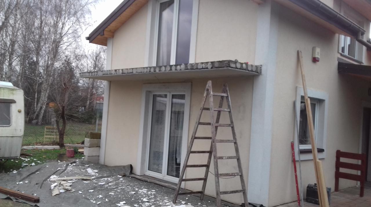 Dwukondygnacyjny dom w trakcie prac remontowych, widoczne okno PCV z ramą, drewniana drabina oparta o ścianę, folia ochronna na ziemi i resztki gruzu, w tle przyczepa kempingowa i drzewa bez liści.
