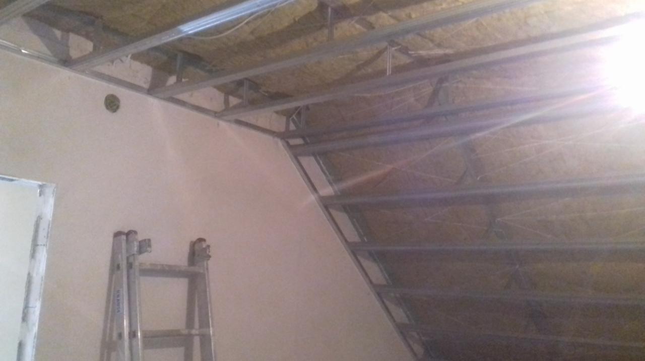 Sufit podwieszany w trakcie montażu: widoczna metalowa konstrukcja, izolacja z wełny mineralnej i fragment ściany z drabinką opartą o nią.