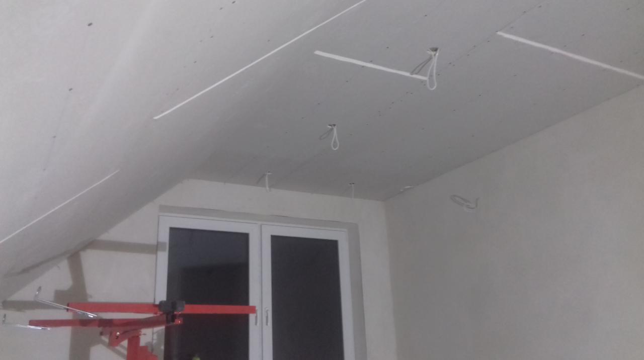 Instalacja sufitu podwieszanego z płyt gipsowo-kartonowych, widoczne profile i uchwyty montażowe, częściowo wykończona powierzchnia, czerwony podnośnik do płyt g-k przy ścianie, okno w tle.