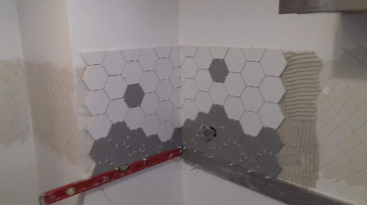 Układanie heksagonalnych płytek w odcieniach szarości i bieli na ścianie w rogu pomieszczenia, widoczny poziomica i nierównomiernie nałożony klej.