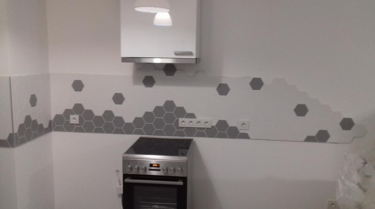 Fragment kuchni z nową aranżacją ściany nad blatem, wykończoną białymi płytkami z szarymi heksagonalnymi akcentami, widoczne gniazdka elektryczne i kuchenka.