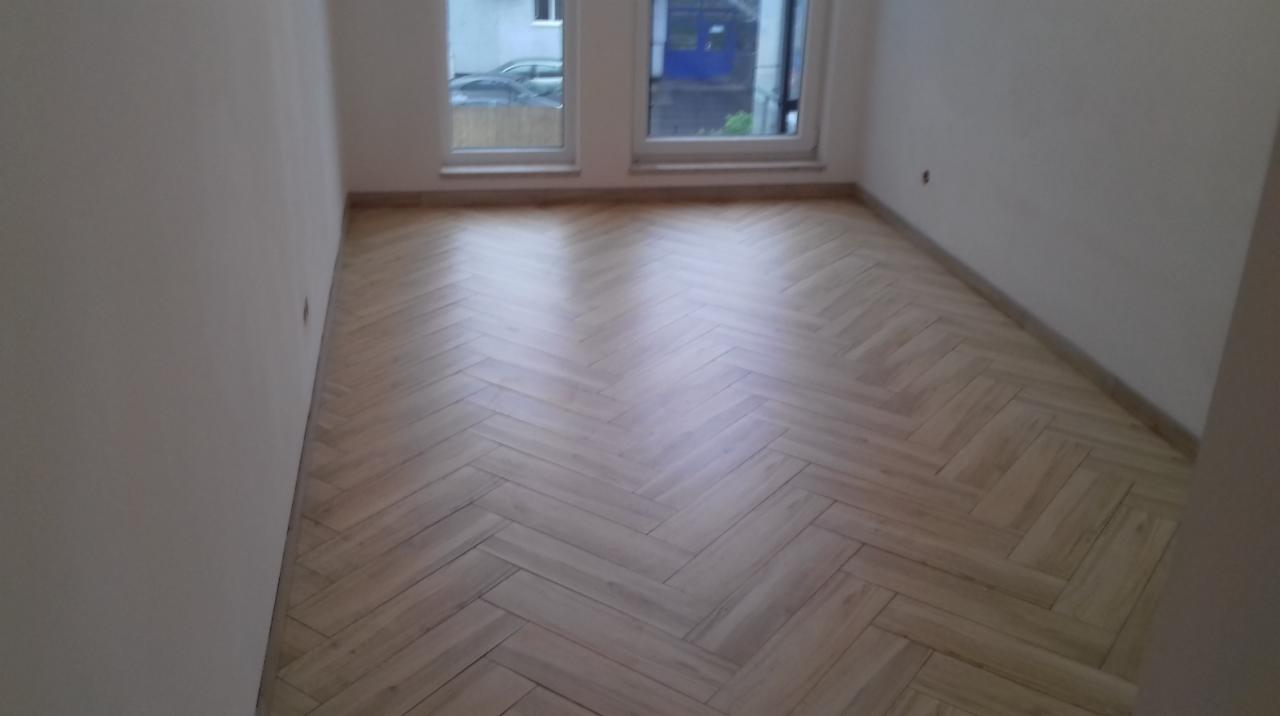 Jasne, drewniane panele ułożone w jodełkę na podłodze w pustym pokoju z dwoma oknami.