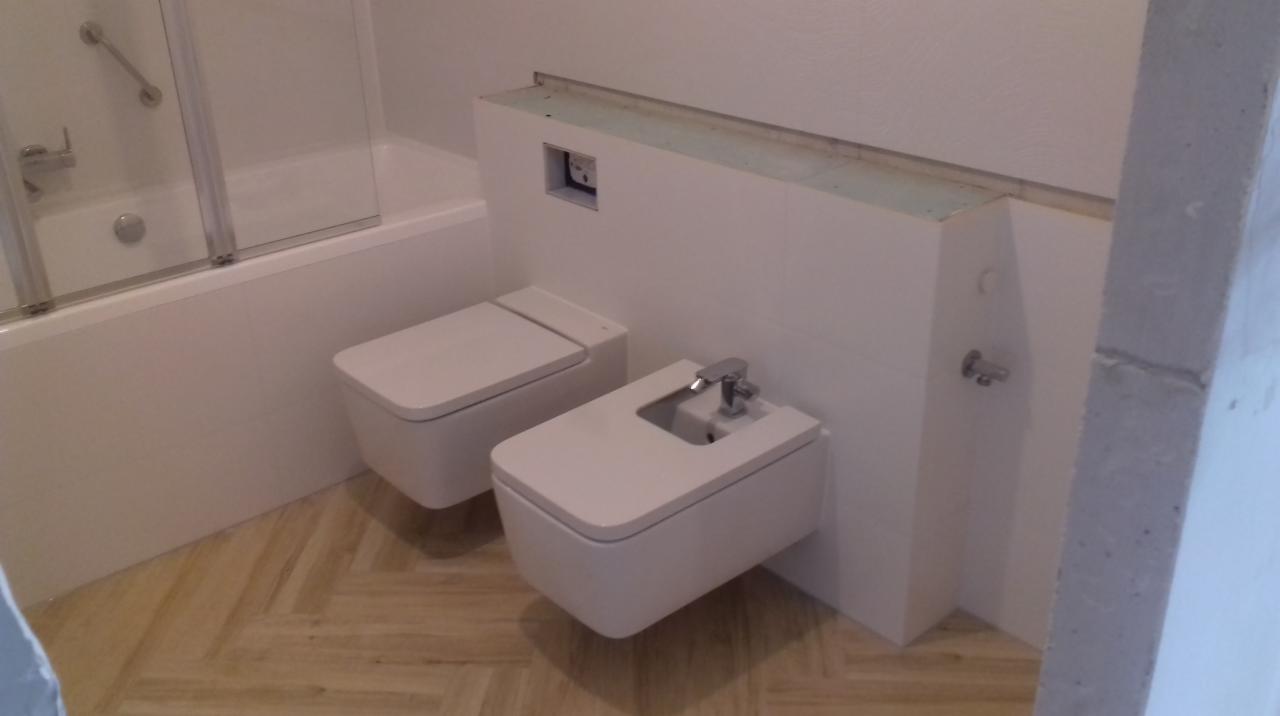 Nowoczesna łazienka z podwieszanym WC i bidetem, wykończona w bieli z drewnianą podłogą w jodełkę. Widoczna instalacja podtynkowa i fragment wanny z parawanem.