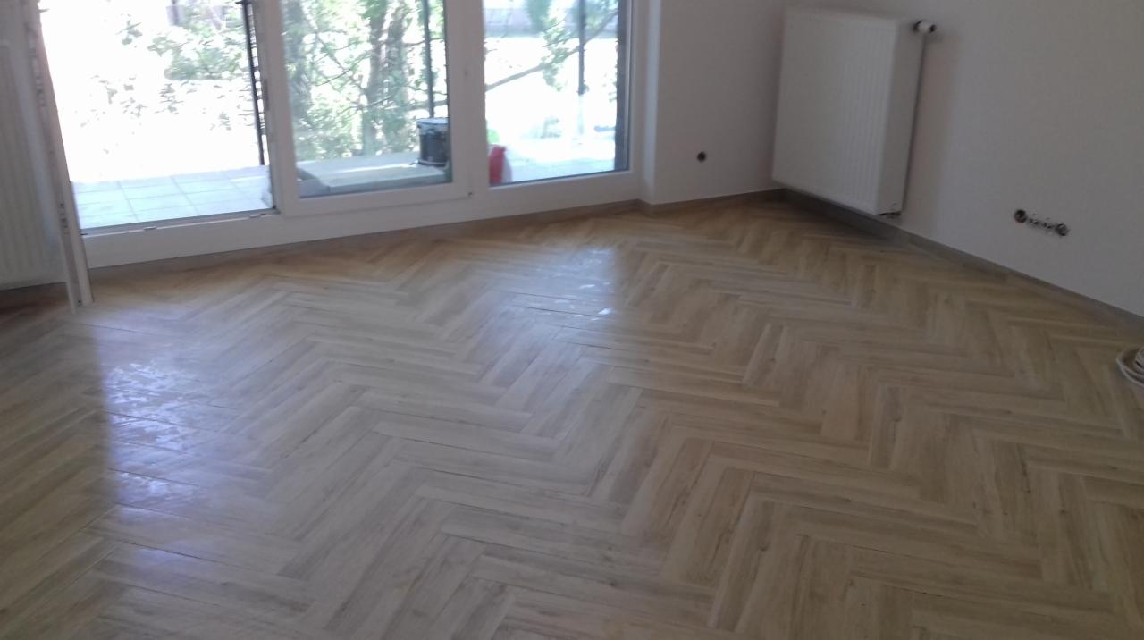 Jasny, odnowiony parkiet ułożony we wzór jodełki w pustym pokoju z dużym oknem balkonowym i grzejnikiem na ścianie.