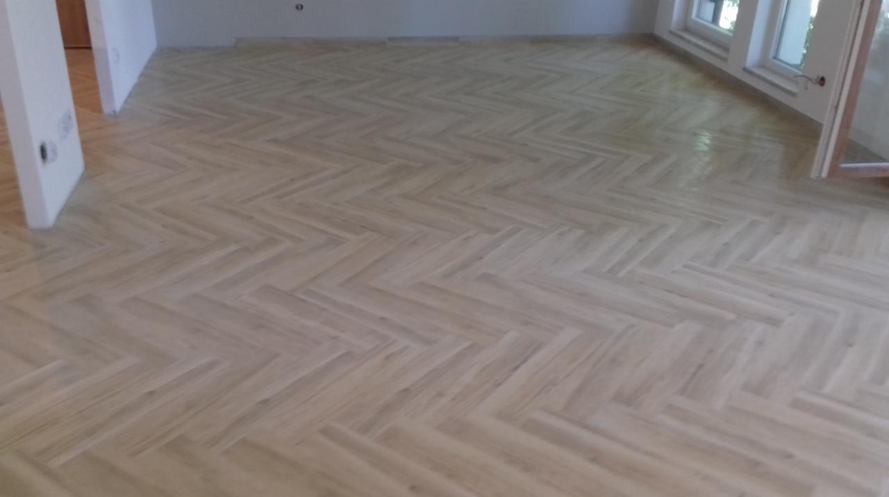 Jasny, parkiet ułożony we wzór jodełki francuskiej, świeżo po lakierowaniu, widoczne odbicie światła, białe listwy przypodłogowe w tle.