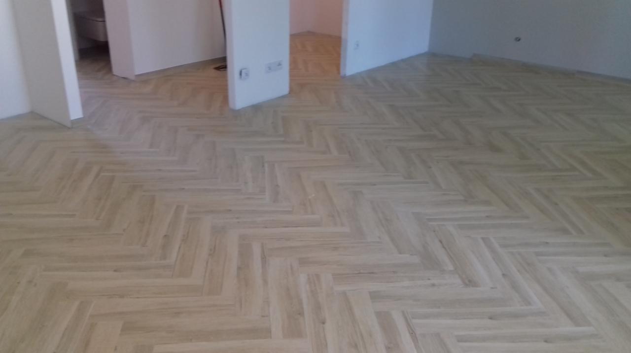 Ułożona pod kątem jasna podłoga panelowa imitująca drewno w pustym pokoju, widoczne gniazdka elektryczne na ścianie i otwarte drzwi do sąsiednich pomieszczeń.